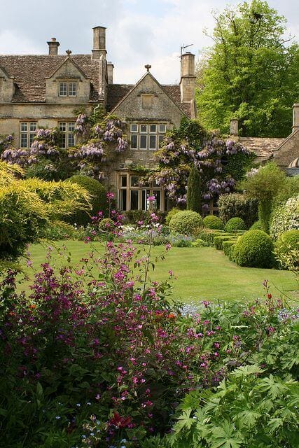 A vibrant DIY garden featuring pink and purple flowering plants, low hedges, and large trees, enhancing a historic stone residence.