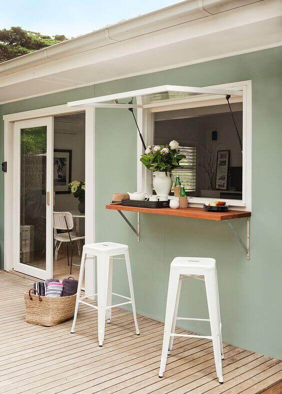 Compact outdoor bar area with a retractable wooden counter, white metal stools, and soft green wall, perfect for back porch ideas.