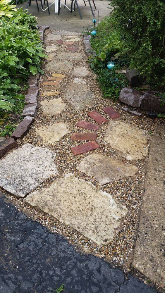 Pathway with irregular flagstones and reddish bricks, surrounded by lush greenery. Gravel base ensures stability and drainage.