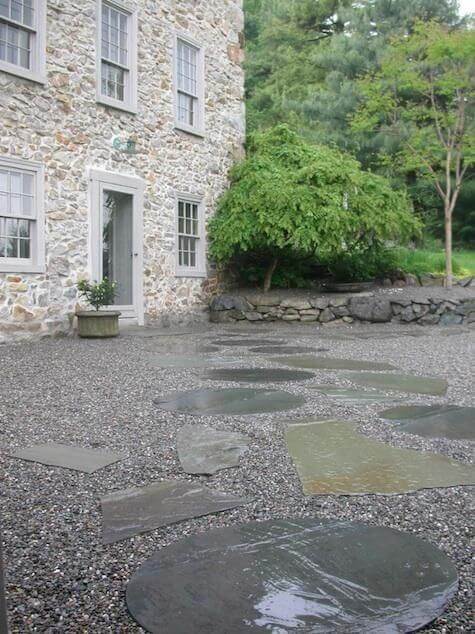 Large slate stones create a flagstone and gravel walkway, leading to a rustic stone structure, surrounded by lush vegetation.