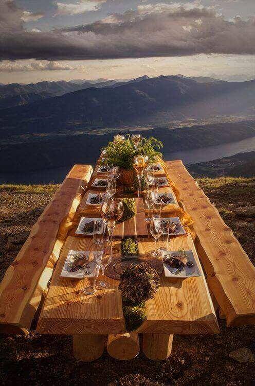 Long, rustic live edge dining table made from large logs, set outdoors with panoramic mountain views, featuring eight elegant place settings.