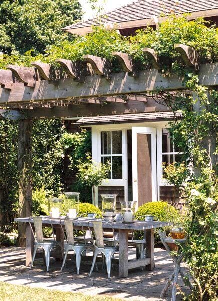 A wooden pergola creates a shaded dining patio, adorned with climbing plants, a reclaimed wood table, and metal chairs, enhancing outdoor gatherings.