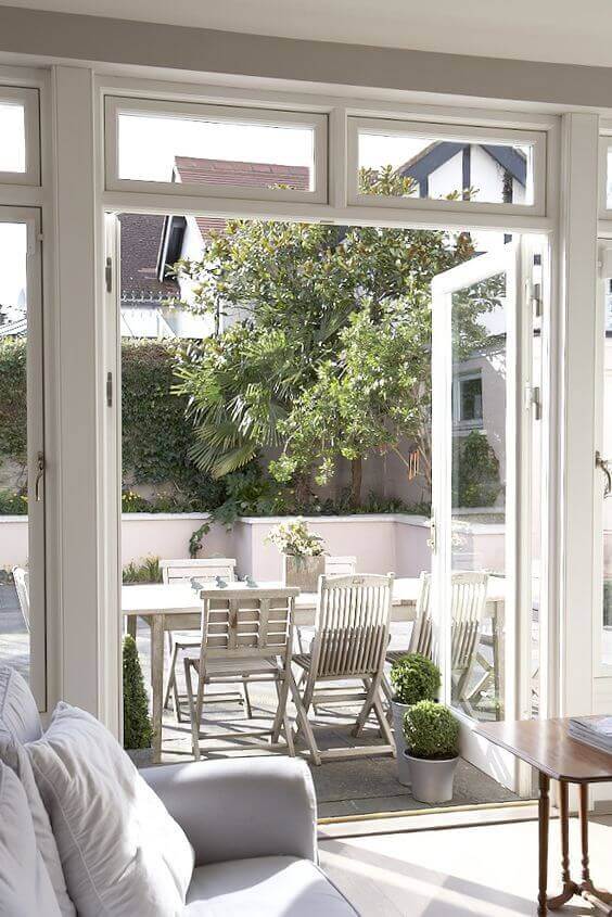 Outdoor dining area with a rectangular wooden table and folding chairs, surrounded by greenery and light-colored stone pavers, ideal for patio door ideas.