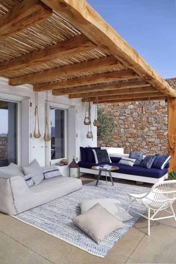 Cozy outdoor living space with a sectional sofa, lounge chair, and wooden pergola. Features a woven rug, coffee table, and decorative lanterns.