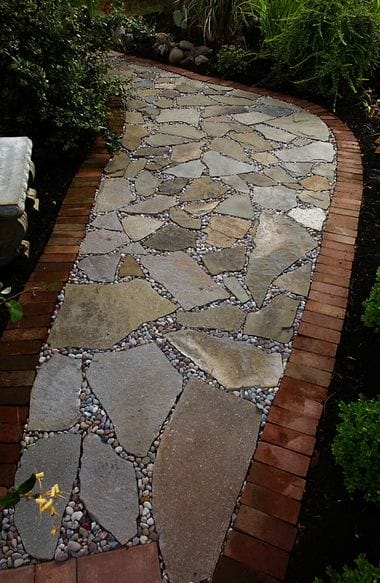 Irregularly shaped flagstone slabs form a 3-foot-wide walkway, bordered by red brick, with gravel and lush shrubs enhancing the garden's appeal.