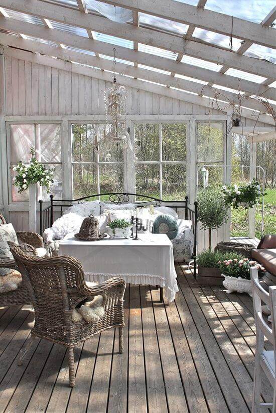 Sunlit back porch features large glass windows, reclaimed wood flooring, wicker seating, and potted plants, creating a cozy, inviting space.