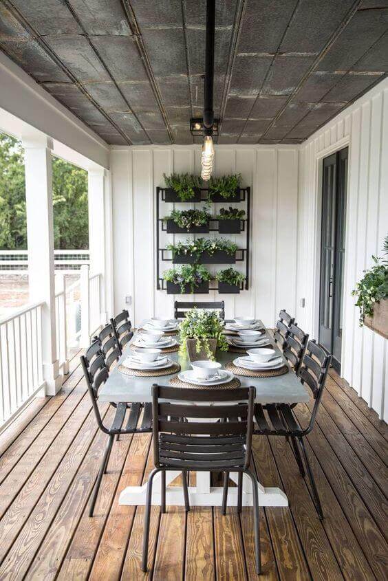 An outdoor dining area with a large concrete table for twelve, surrounded by black metal chairs, on a warm-toned wooden deck.
