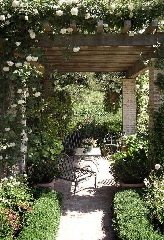 Structured outdoor sitting area under a wooden pergola, adorned with climbing white roses, featuring wrought iron furniture and laid brick pathways.