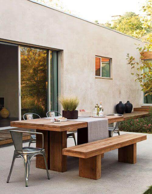 Substantial live edge dining table made of walnut, flanked by rustic benches and metal chairs, set in a warm outdoor area with greenery.