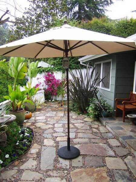 Patio umbrella shaded by tropical plants, alongside a flagstone and gravel walkway leading to a residential structure.