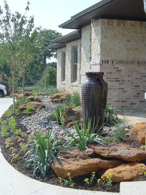 Striking front yard landscape with textured boulders, smooth river stones, a tall ceramic vase, and lush flowering perennials, enhancing curb appeal.