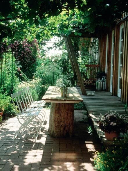 Live edge dining table made from natural wood, supported by a tree stump, surrounded by greenery and white metal chairs for six.
