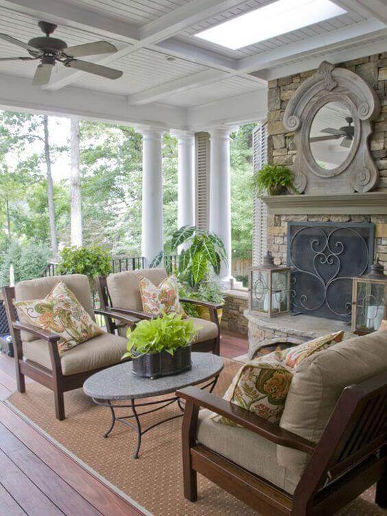 Spacious back porch with floral-patterned armchairs around a stone coffee table, featuring a stone fireplace and skylights, surrounded by greenery.