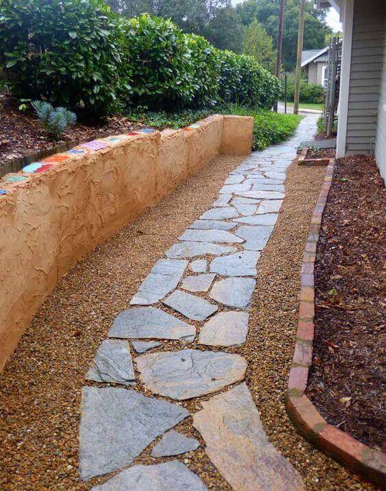 Flagstone and gravel walkway features irregular stone slabs set in fine gravel, bordered by a textured earthen wall and lush greenery.