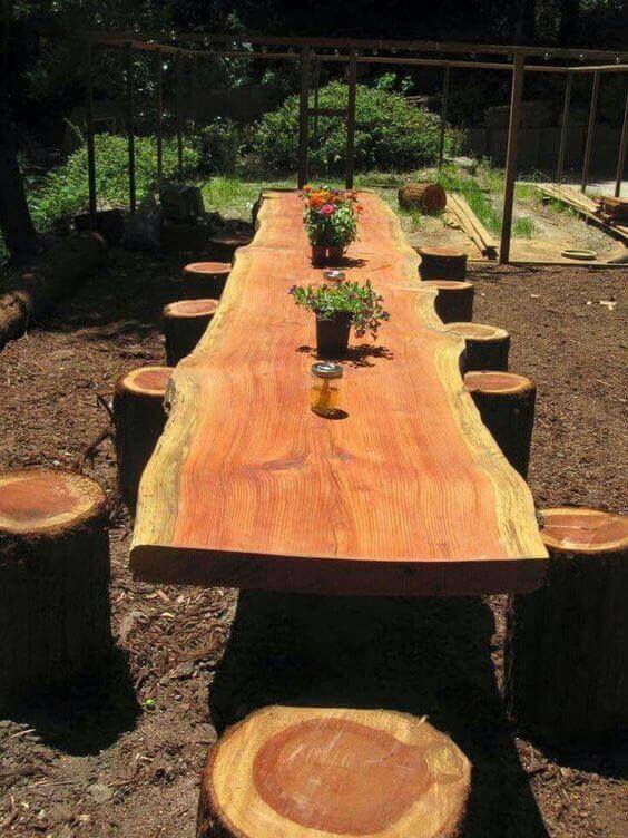 Large live edge dining table made from a single wood slab, surrounded by eight rustic log stools, set in a sunlit garden.