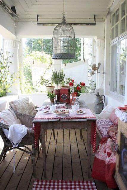 Cozy outdoor dining space with a wooden table, checkered tablecloth, wicker chairs, and a decorative birdcage, surrounded by greenery.