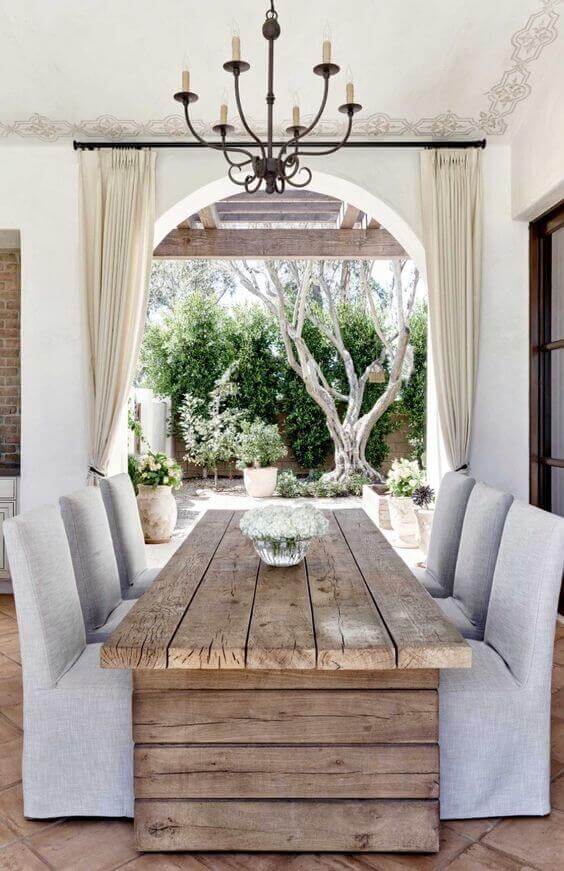 Rustic live edge dining table made from reclaimed timber, surrounded by six light grey upholstered chairs in a serene garden setting.