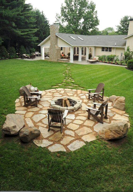 Circular stone patio with a fire pit, surrounded by flagstone pavers and Adirondack chairs. A landscaped flagstone and gravel walkway leads to the house.