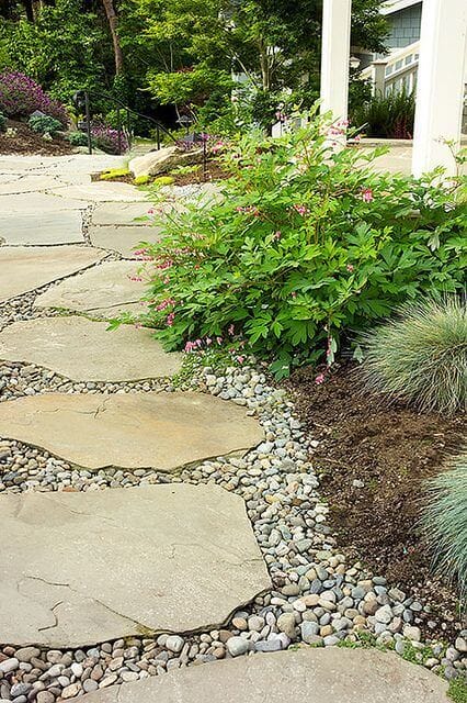 Flagstone pathway bordered by fine gravel and lush greenery, leading through a vibrant garden with flowering shrubs.