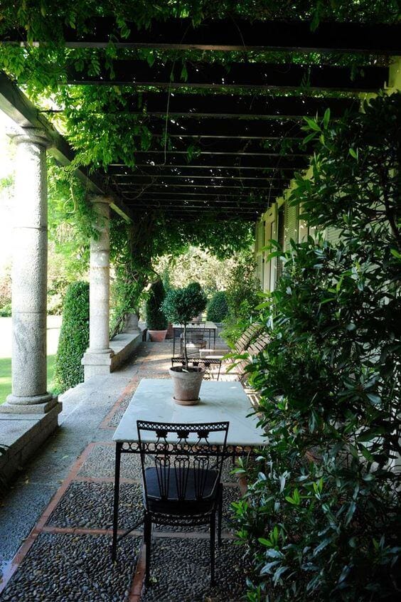 Patio garden with a stone table and wrought iron chairs, featuring a pergola with climbing vines and neatly clipped hedges.