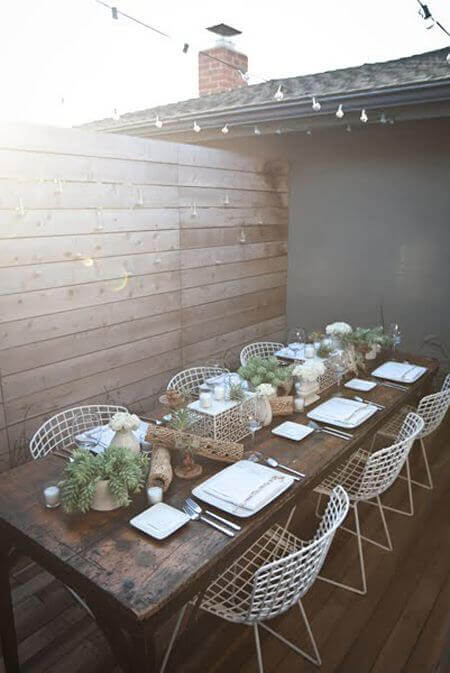 Long wooden dining table made from reclaimed timber, surrounded by twelve white wire chairs, adorned with greenery and ambient string lights.