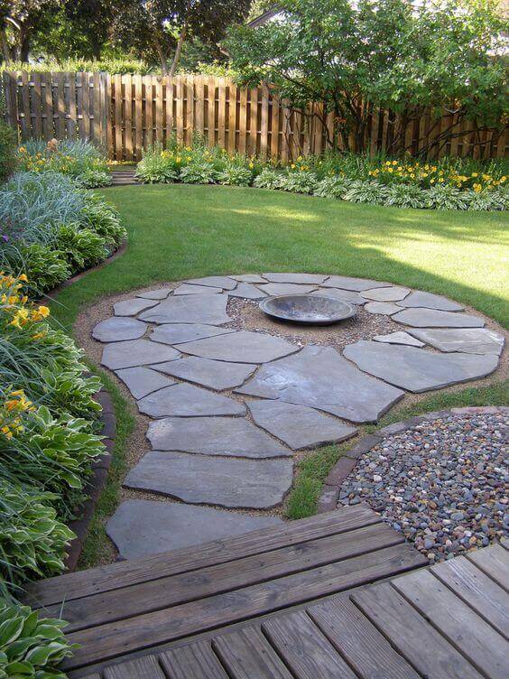 Circular patio features flat flagstone slabs and gravel, with a central fire pit, surrounded by grass and perennial flower beds.