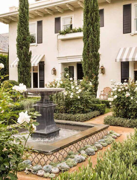Fountain at garden's center, surrounded by succulents and rose bushes, framed by evergreen trees and a gravel pathway, creating a serene landscape.