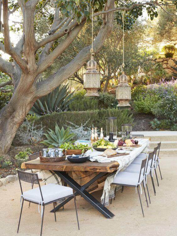 Outdoor dining setup features a live edge dining table made from reclaimed wood, surrounded by metal chairs with gray cushions under a mature tree.