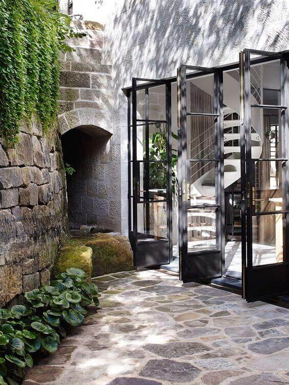 A stone pathway leads to a glass facade with large black-framed doors, featuring a spiral staircase and lush garden foliage.