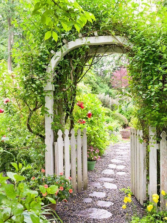 Rustic archway entwined with green vines leads through a vibrant garden pathway, bordered by a white picket fence, ideal for DIY trellis ideas.