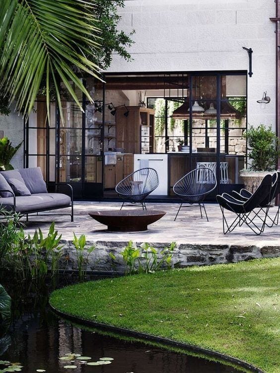 Outdoor living area features a modern stone patio with a low gray sofa, wire-frame chairs, and a glass wall, enhancing indoor-outdoor flow.