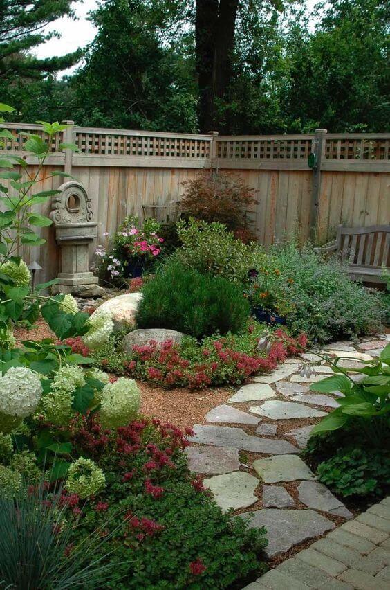 A flagstone and gravel walkway winds through a lush garden, bordered by flowering shrubs and a wooden fence, enhancing the serene landscape.