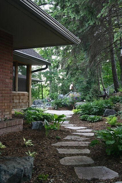 Stone pathway leads through a lush garden, featuring irregular flagstone slabs over mulch, surrounded by diverse plants and ornamental rocks.