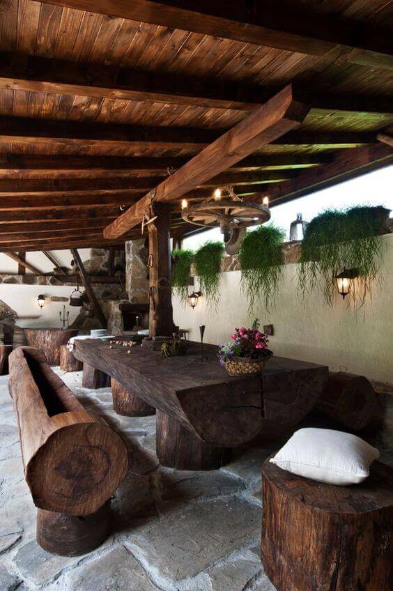 A handcrafted live edge dining table made from thick, polished logs under a rustic pergola, surrounded by log stools and lush ferns.