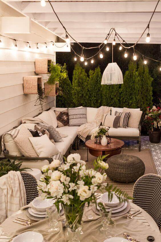 Cozy back porch with a sectional sofa in neutral tones, decorative pillows, circular coffee table, and ambient string lights, surrounded by greenery.