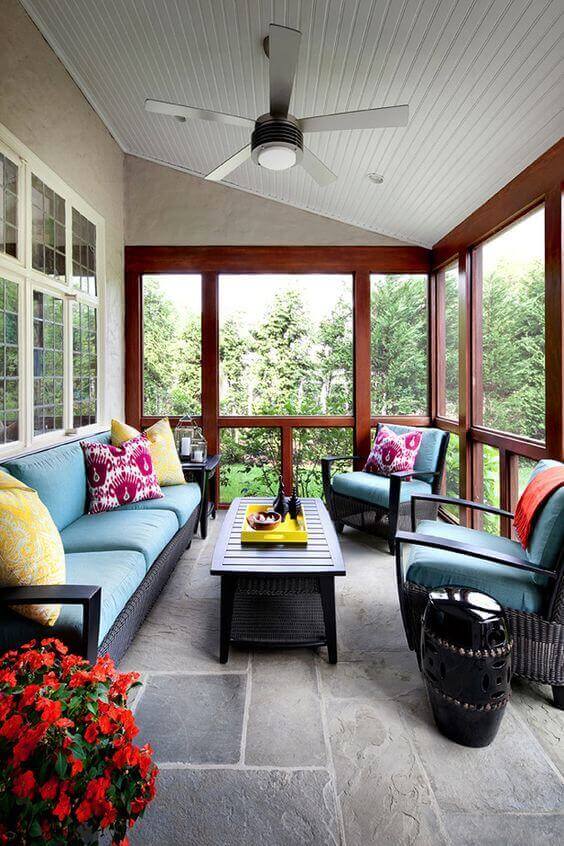Back porch ideas showcase a sunroom with stone flooring, soft blue hues, vibrant seating, and large screened windows for natural light.