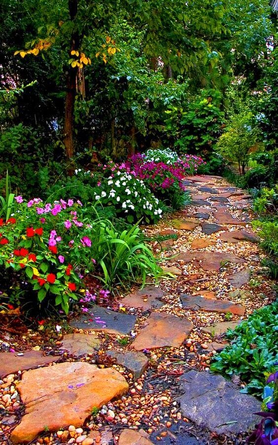Winding flagstone walkway surrounded by vibrant flower beds, featuring pink, white, and red blooms in a lush, shaded landscape.
