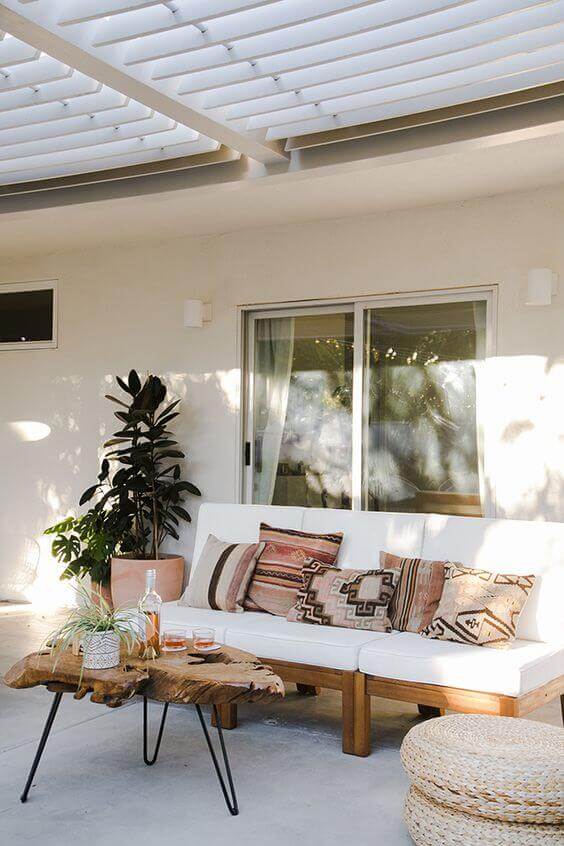 Cozy outdoor living space with a white sofa, organic-shaped wood coffee table, and potted plant, sheltered by a slatted pergola.