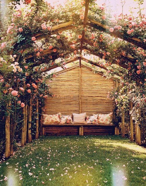 Wooden garden shelter with peaked roof, side trellises featuring pink climbing roses, cozy bench, and lush green grass, perfect for relaxation.
