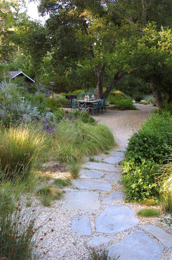 Lush garden pathway with flagstone and gravel, featuring a rustic dining area amid mature trees and vibrant perennials.