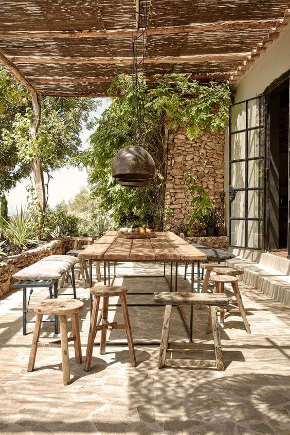 Live edge dining table in a rustic outdoor setting, featuring a 2.5m reclaimed wood surface, black metal legs, and wooden stools amidst lush greenery.