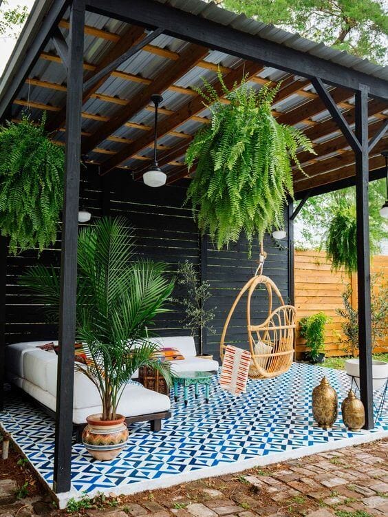 Cozy outdoor living space with a white sofa and hanging chair under a canopy, featuring a geometric blue tiled floor and lush greenery.