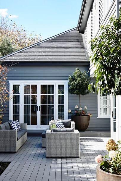 Contemporary outdoor seating area with gray wicker sofas on a composite deck, bordered by a gray house and green foliage, ideal for relaxation.
