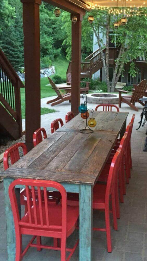 Live edge dining table in a rustic outdoor area, surrounded by greenery, with 10 red chairs under string lights on a covered porch.