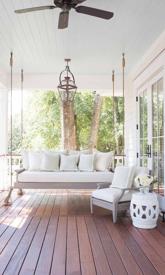 Outdoor living space with a suspended daybed on rich mahogany decking, light cushions, ceiling fan, and lantern-style lighting. Two mature trees enhance the view.