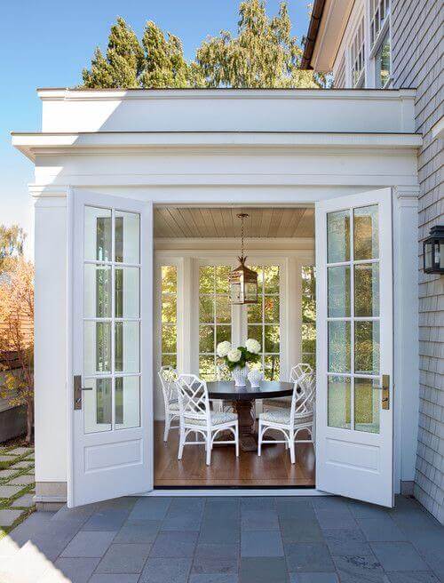 Sunlit patio featuring large glass doors leading to a landscaped garden, with a circular wooden dining table and six white wicker chairs.