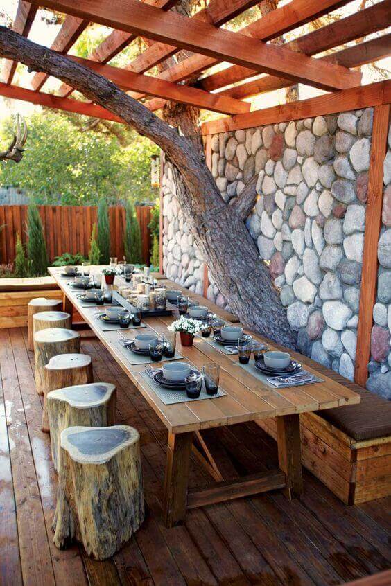 Rustic outdoor dining area with a live edge dining table made from reclaimed timber, surrounded by wooden stump stools and a natural stone wall.