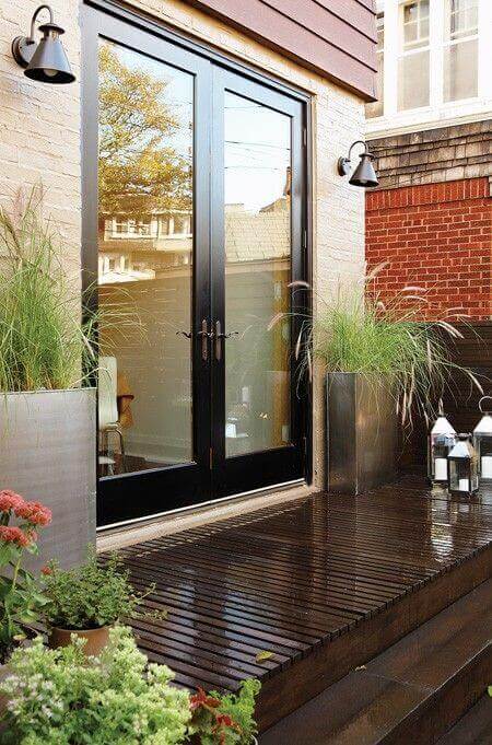 Sleek wooden deck with large glass doors leading to an outdoor living space, flanked by stainless steel planters with ornamental grasses.