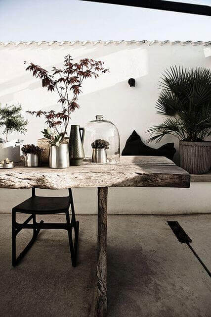 Rustic live edge dining table made from reclaimed timber, featuring a pedestal base and surrounded by modern black chairs and potted plants.