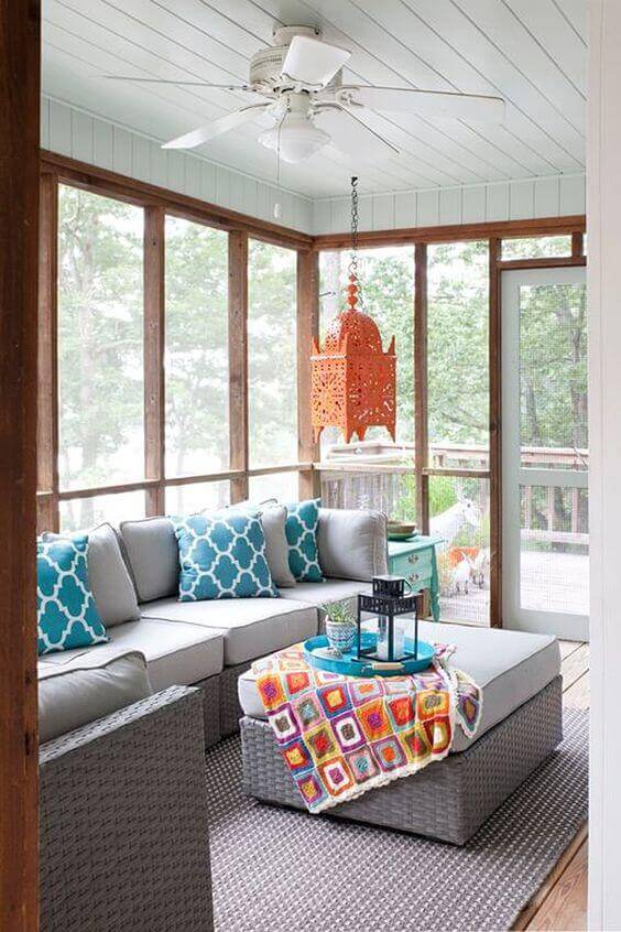 Screened-in back porch with a sectional sofa in turquoise and white, vibrant blanket on a central table, and large windows for outdoor views.